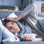 Sick exhausted girl in scarf is lying in bed wrapped in blanket. Young woman with fever and headache is measuring temperature with thermometer, treated at home. Winter cold and flu concept.