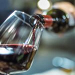 person holding a bottle of red wine, is pouring some red wine in a wine glass.