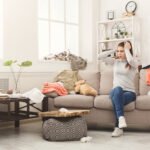 Desperate helpless woman sitting on sofa in messy living room. Young girl surrounded by many stack of clothes. Disorder and mess at home, copy space